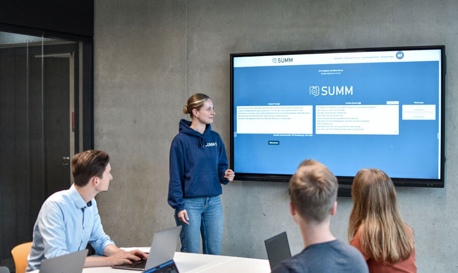 Here are people in a room. There is a big TV in the room. On the screen of the TV you can see the website and the translation tool of SUMM. Three people are sitting at the table and looking at the TV. A woman stands to the left of the TV and looks at it as well. All the people are having a meeting.