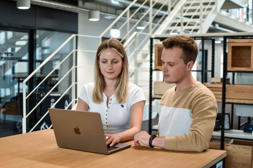 A woman and a man are standing at a table. There is a laptop on the table. Both are looking at the laptop and reading a blog post. The woman is typing something. We are looking at the two people from the front here. Therefore, you can't see the screen of the laptop.