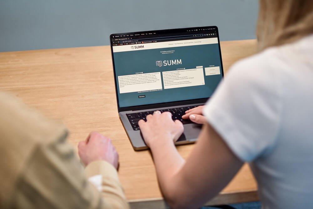 summ-uebersetzer-tool Zwei Menschen sitzen an einem Tisch. Auf dem Tisch ist ein Laptop. Ein Mensch tippt auf dem Laptop etwas ein. Der Laptop zeigt die Webseite von SUMM und das Übersetzungstool. Wir blicken hier von hinten wie die zwei Menschen auf den Laptop.