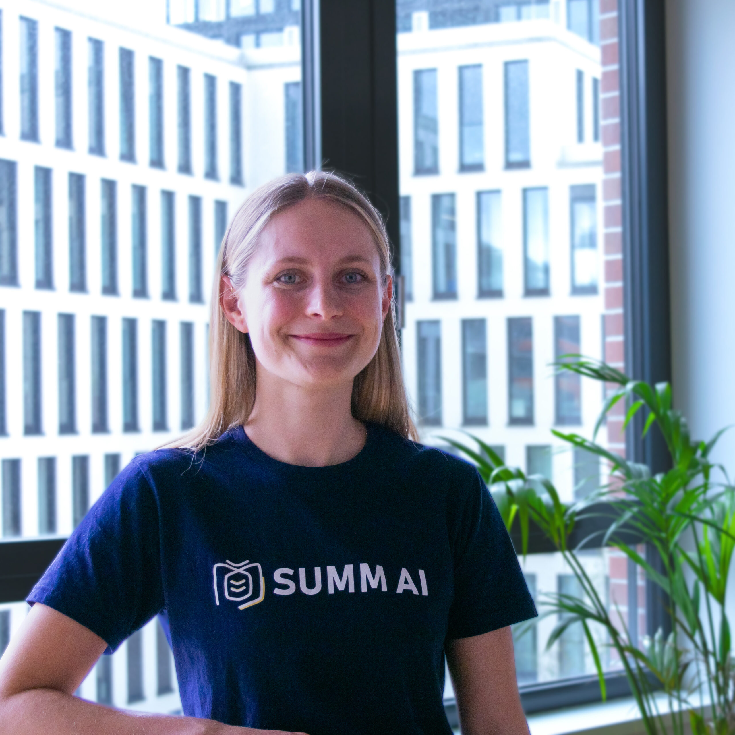 Ein Bild unserer Gründerin Vanessa. Sie steht im Coworking-Bereich des Werk1, lehnt mit einem Arm auf einem Tisch und blickt in die Kamera. Sie trägt ein T-Shirt mit dem Logo von SUMM AI.