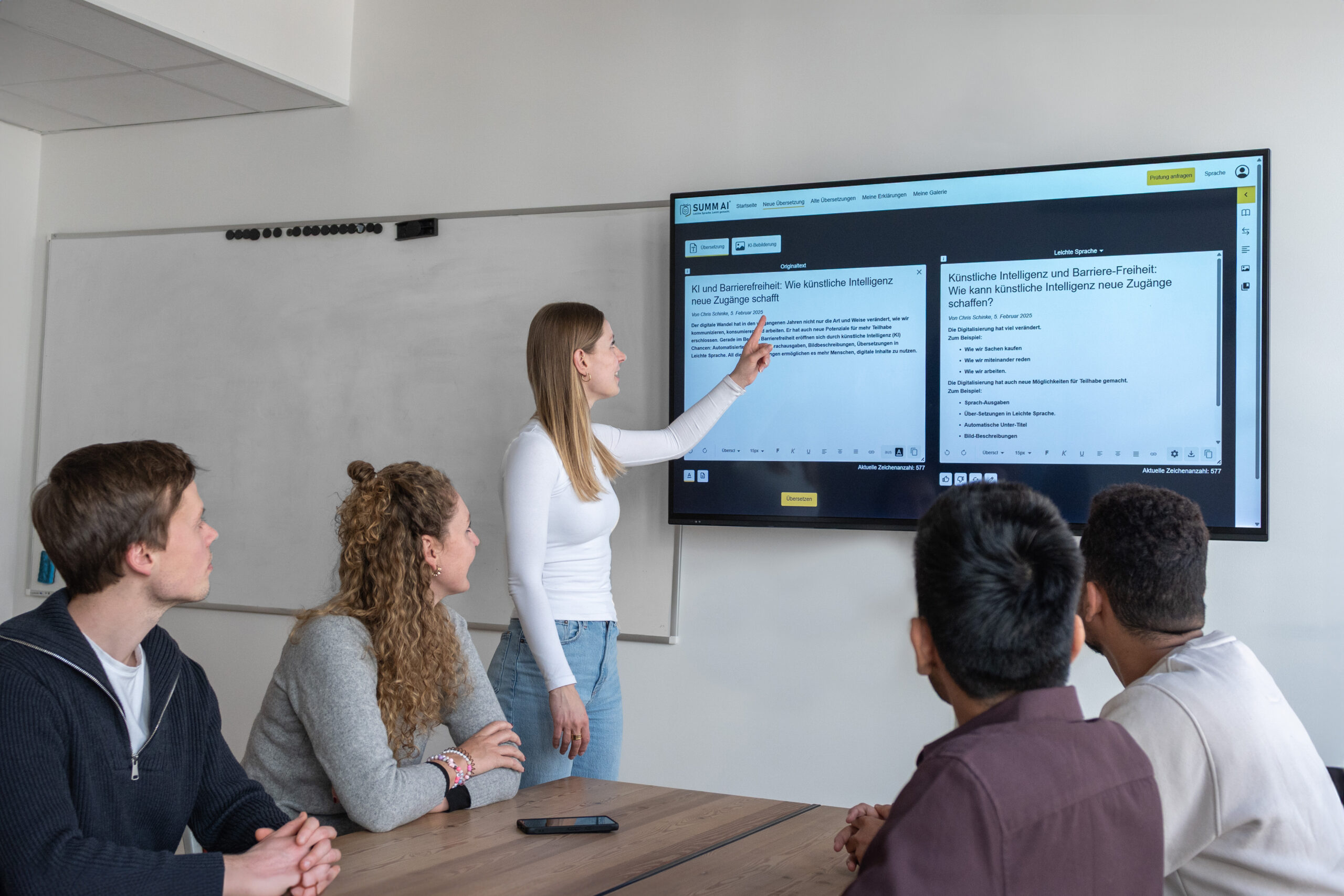 Präsentation Eine der Geschäftsführerinnen von SUMM AI steht vor einer digitalen Tafel und erklärt vier an einem Tisch sitzenden Mitarbeiter:innen etwas.