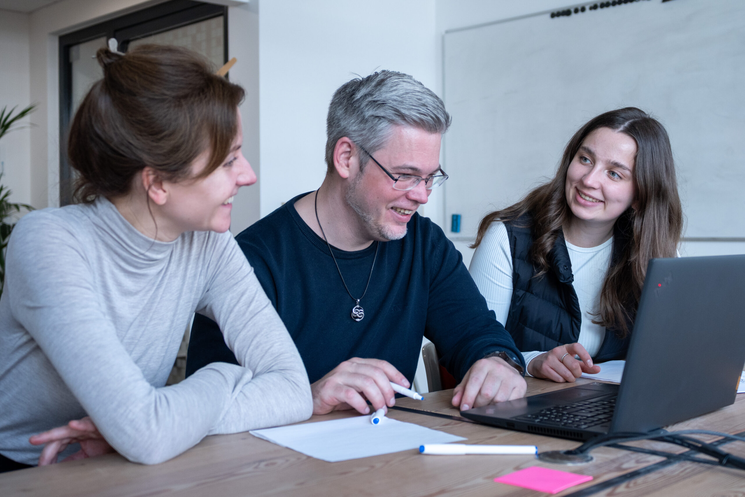 Foto Teamwork Zwei Mitarbeiterinnen und ein Mitarbeiter von SUMM AI sitzen nebeneinander in den Büroräumen an einem Tisch, schauen gemeinsam in einen aufgeklappten Laptop und reden miteinander.
