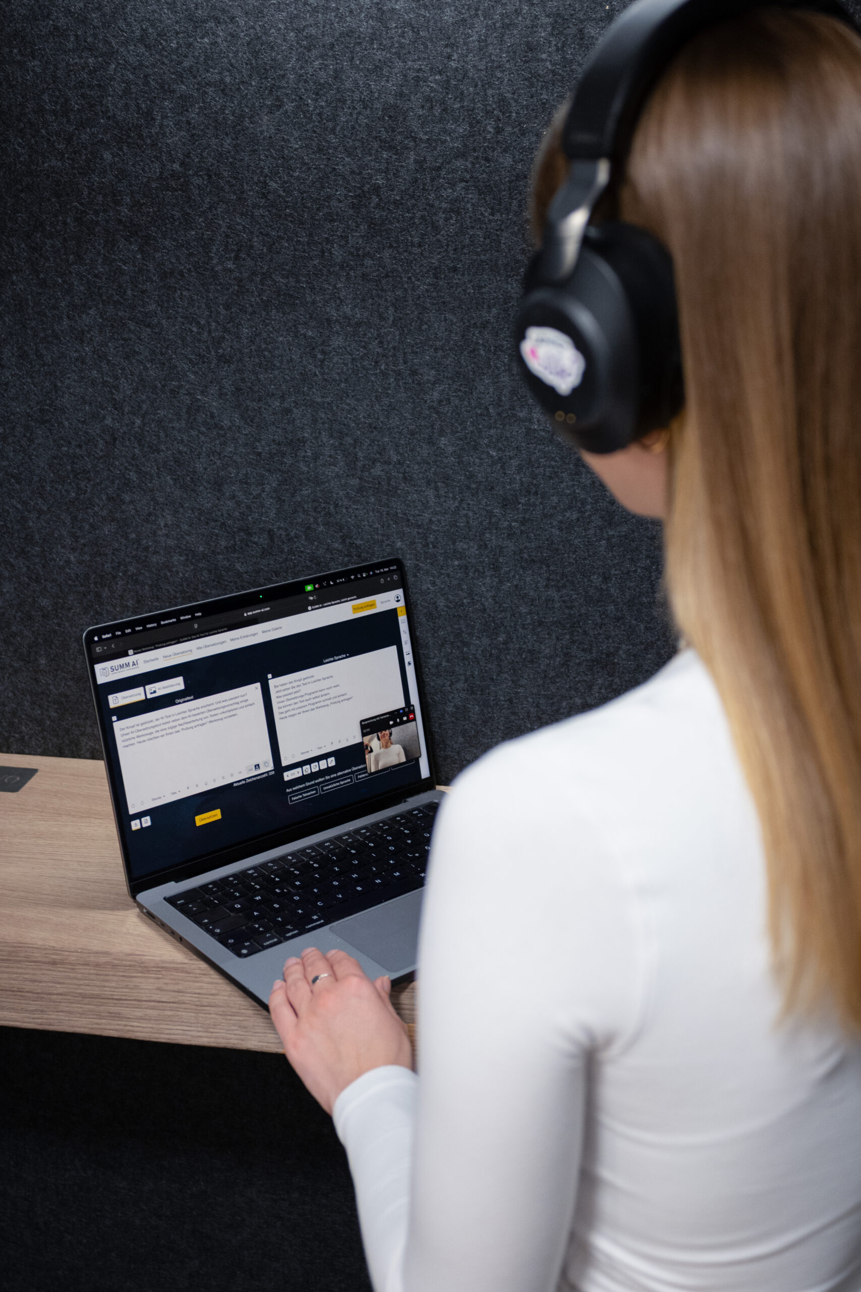 Frau mit langen braunen Haaren, von hinten aufgenommen, sitzt an einem Holztisch vor einem Laptop. Sie trägt einen weißen langärmeligen Pullover und große schwarze Kopfhörer. Auf dem Laptopbildschirm ist eine Webseite mit der Überschrift „SUMM AI“ zu sehen, auf der zwei Textfelder angezeigt werden, vermutlich eine Übersetzungs- oder Textbearbeitungssoftware. Daneben ist ein kleines Video-Chat-Fenster geöffnet. Die Umgebung wirkt modern und minimalistisch, mit dunkler Wand im Hintergrund.