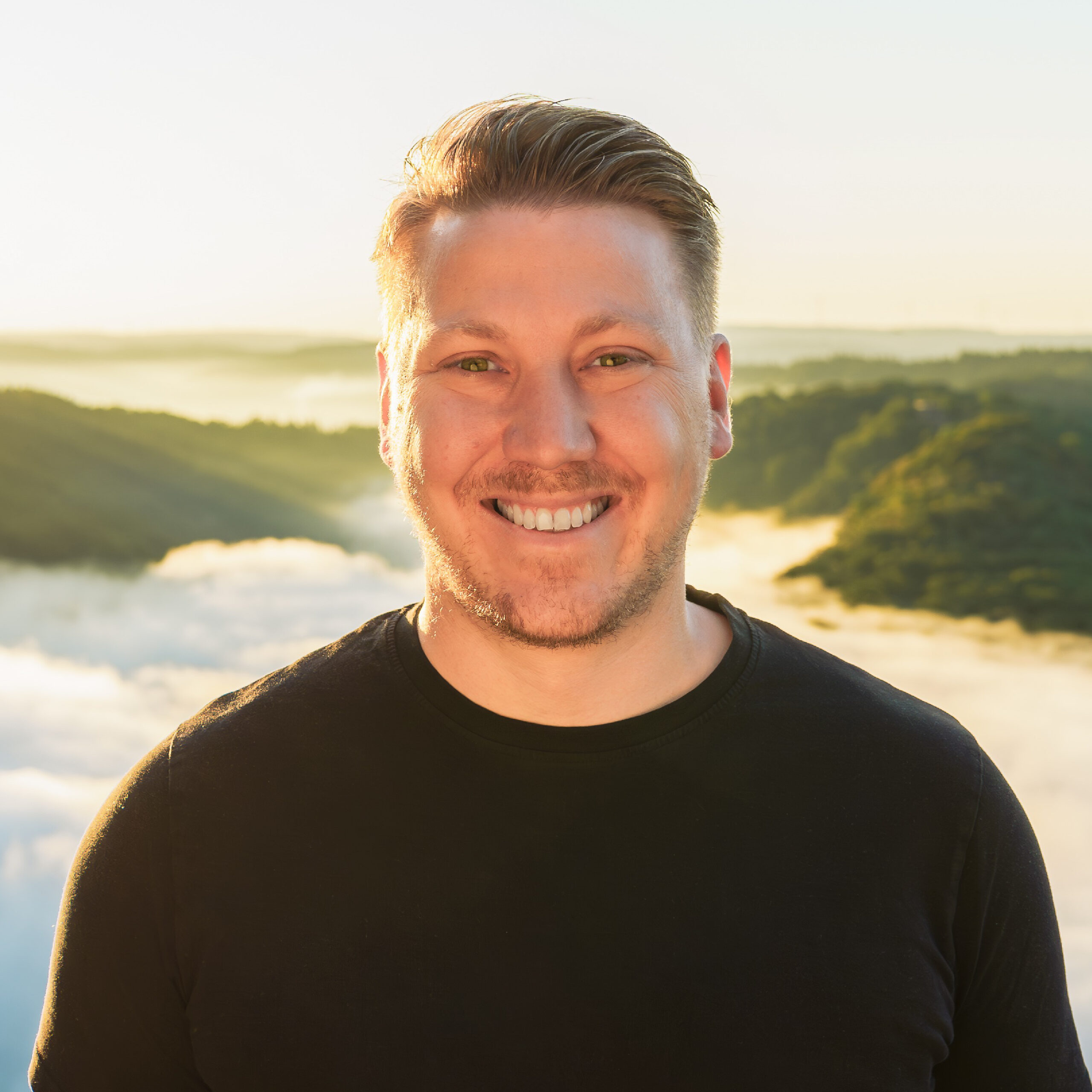 Ein junger Mann mit braunen kurzen Haaren und kurzem Bart, der lachend in die Kamera blickt.
