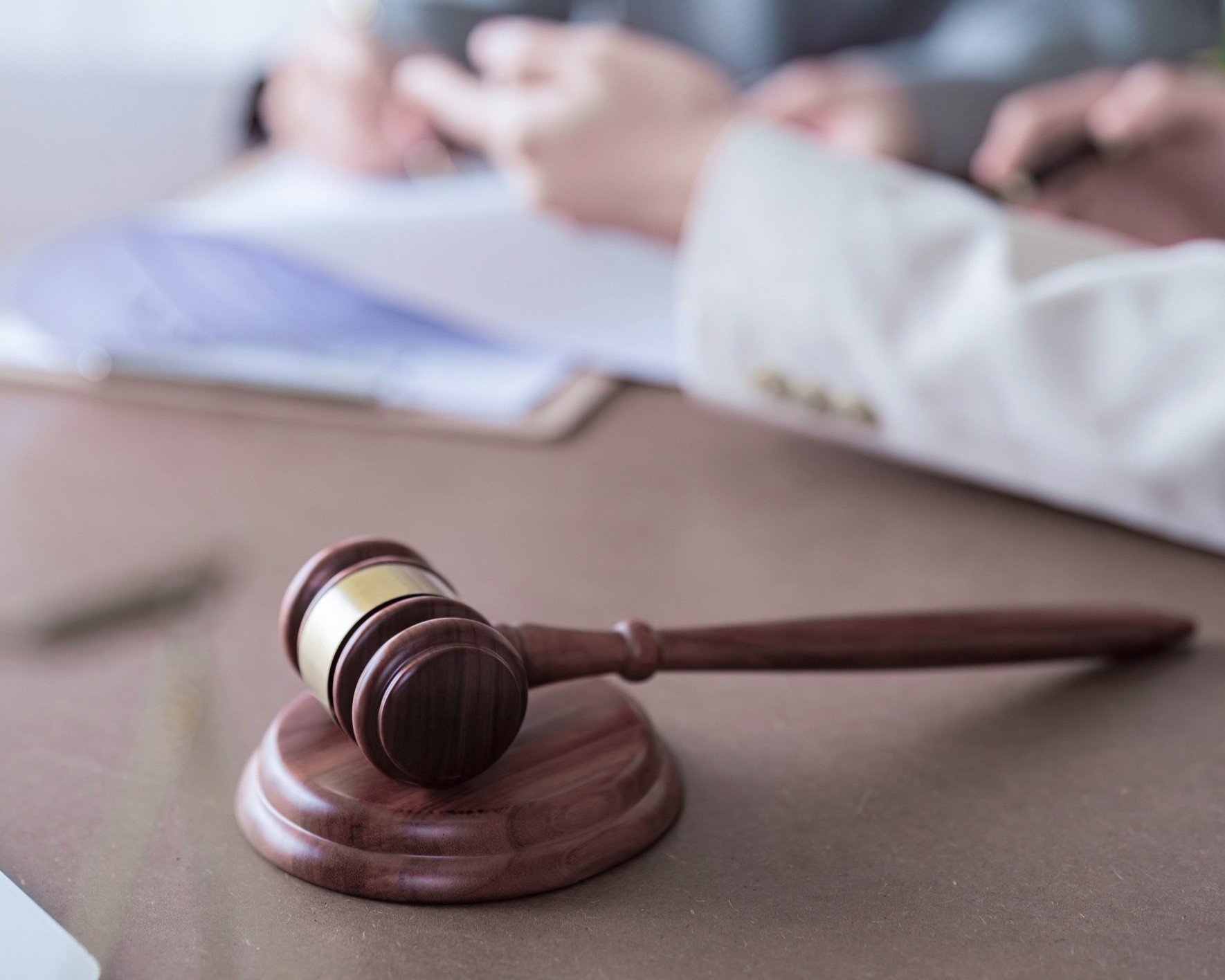 Gerichtsverfahren Ein hölzerner Richterhammer liegt auf einem Tisch im Vordergrund. Im Hintergrund sitzen unscharf zwei Personen in formeller Kleidung, die über Dokumente sprechen oder unterzeichnen – eine Szene, die auf juristische Beratung, ein Gerichtsverfahren oder eine rechtliche Besprechung hinweist.