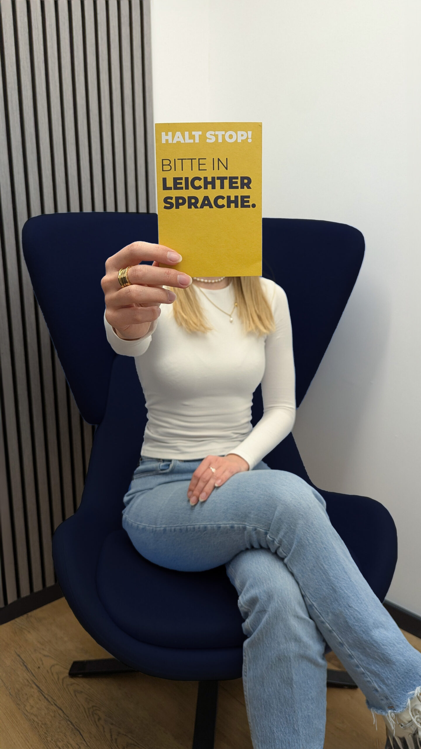 Eine Frau sitzt auf einem dunkelblauen Sessel und hält eine gelbe Karte vor ihr Gesicht. Auf der Karte steht in großen Buchstaben: „HALT STOP! BITTE IN LEICHTER SPRACHE.“ Der Hintergrund ist schlicht mit einer hellen Wand und vertikalen Holzlamellen.