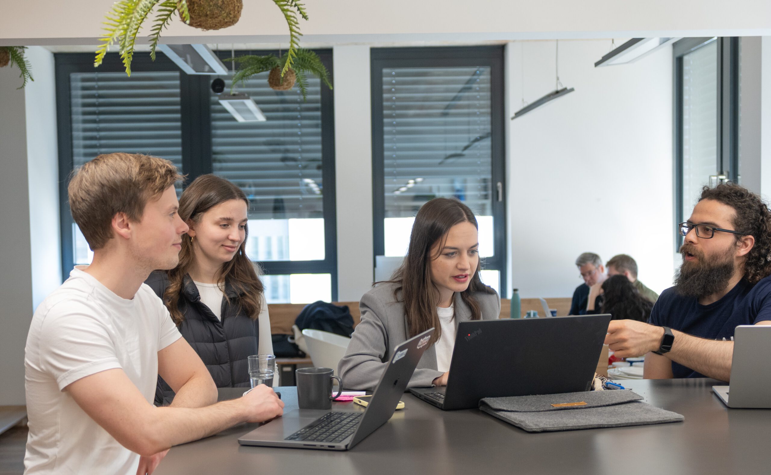 Eine Frau und ein Mann stehen an einem Tisch. Auf dem Tisch steht ein Laptop. Beide schauen auf den Laptop. Die Frau tippt etwas ein. Wir blicken hier von vorne auf die zwei Menschen. Daher sieht man den Bildschirm vom Laptop nicht.